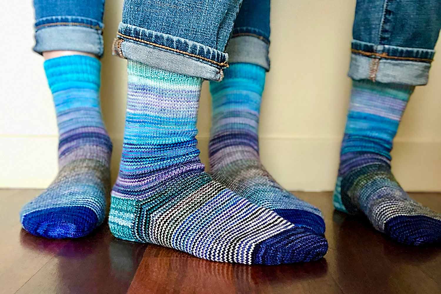 Two pairs of feet wearing one-row striped socks in a fade of colours with solid colour cuffs, heels, and toes.