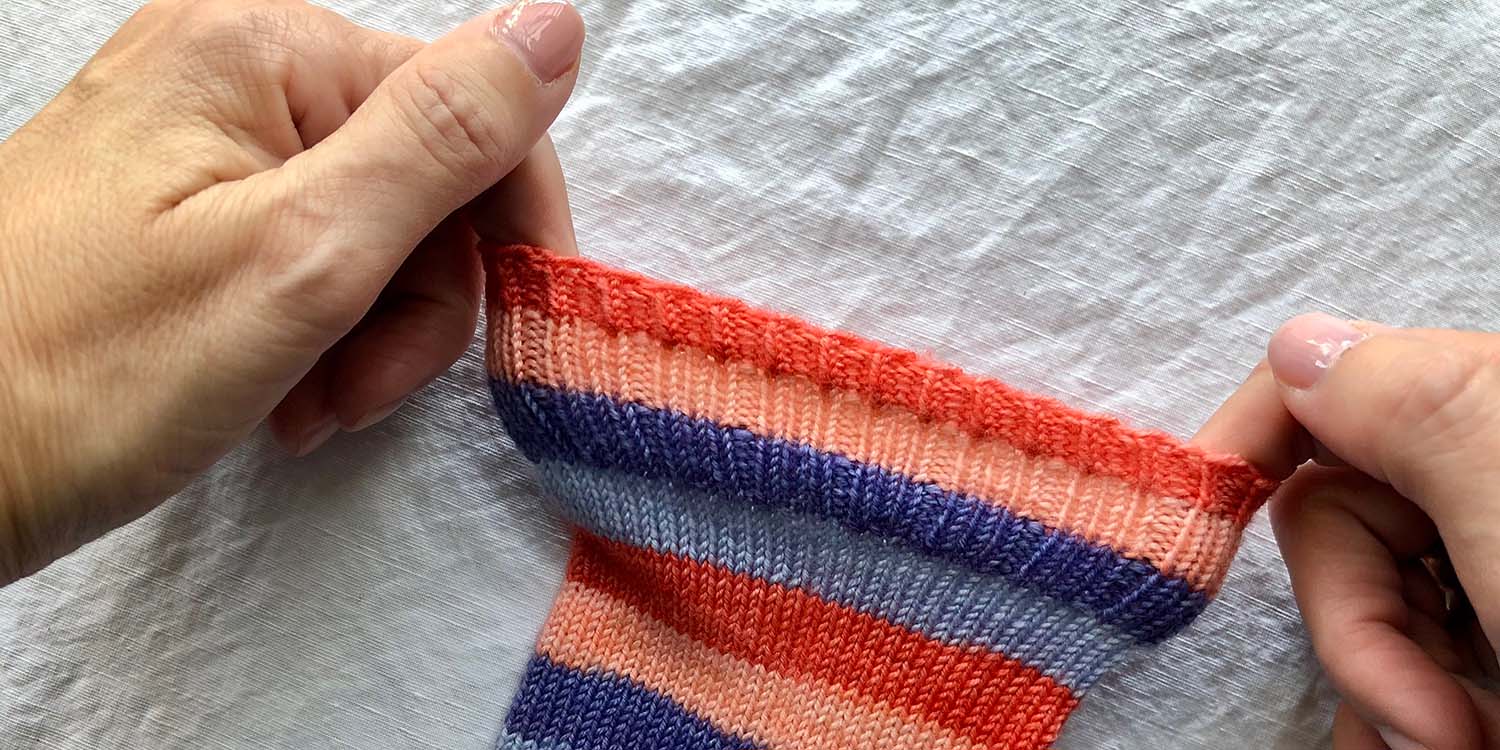 Two hands stretching out top edge of cuff on striped sock, demonstrating how stretchy it is.
