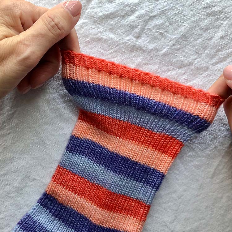 Two hands stretching out top edge of cuff on striped sock, demonstrating how stretchy the bind off is.