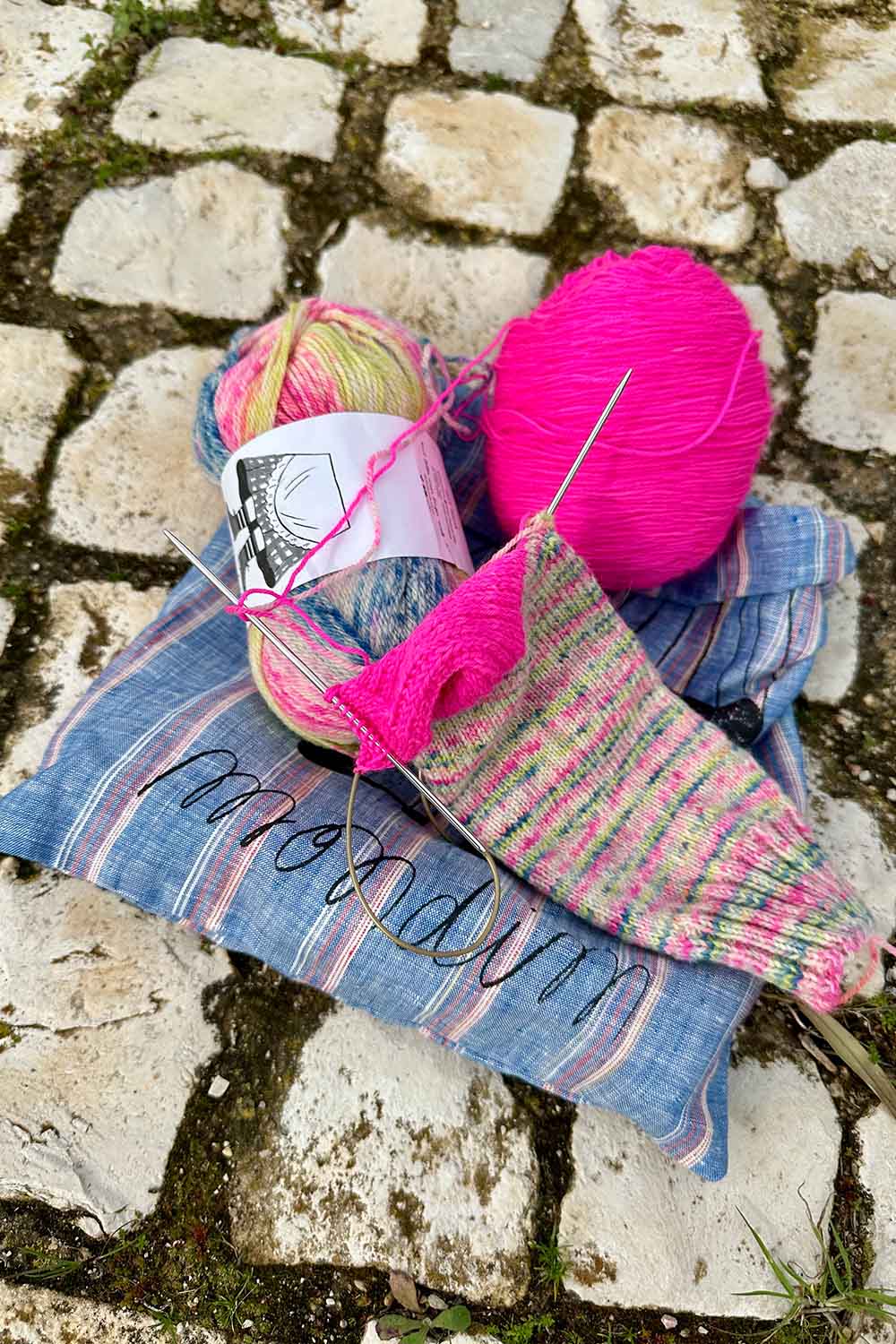 Partially knit sock and two contrasting balls of yarn sitting on project back on top of cobblestone sidewalk.