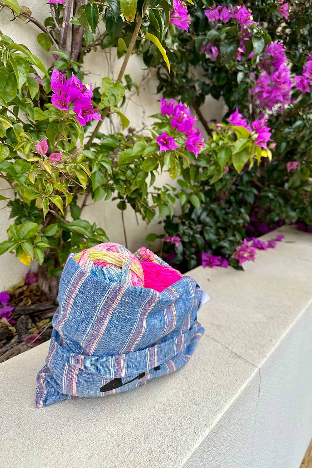 Knitting project bag sitting on a white wall in front of hot pink bougainvillia.