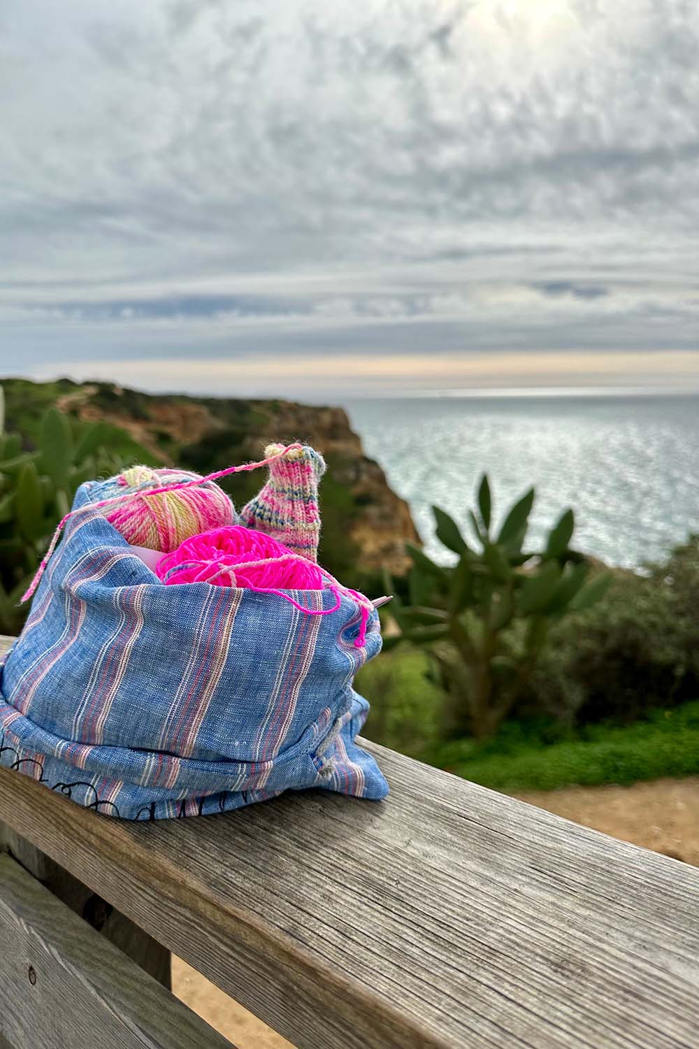 Knitting project bag sitting on a wooden railing with view of ocean and cliff in background.