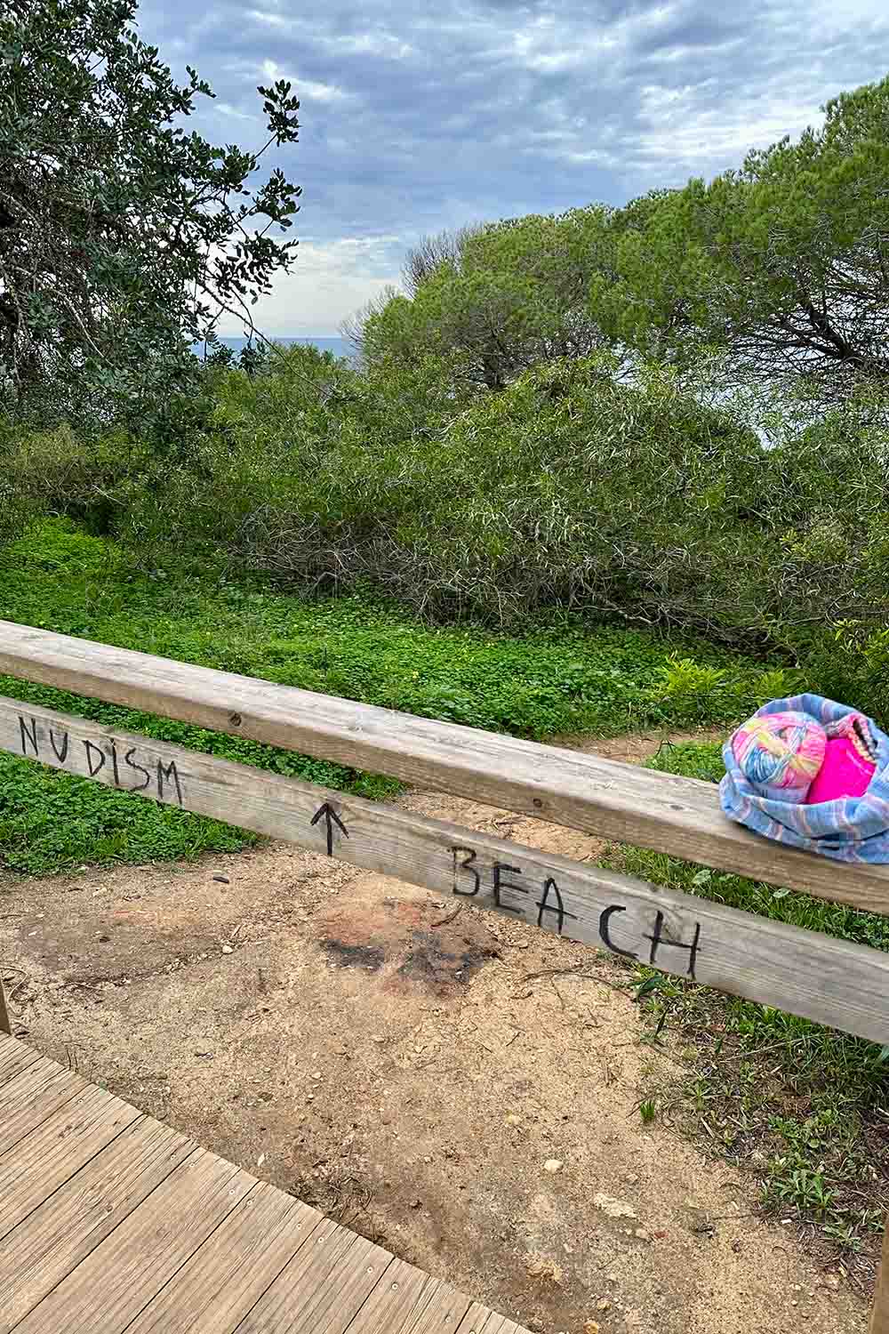Knitting project bag sitting on wooden railing; Nudism Beach and an arrow is hand written on wood slat.