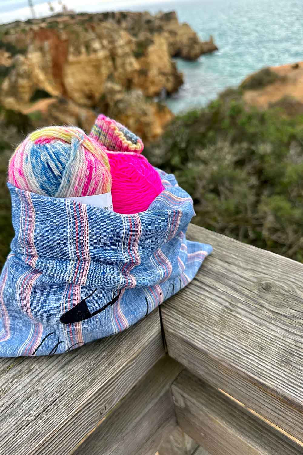 Knitting project bag sitting on a wooden railing with view of ocean and rock formations in background.