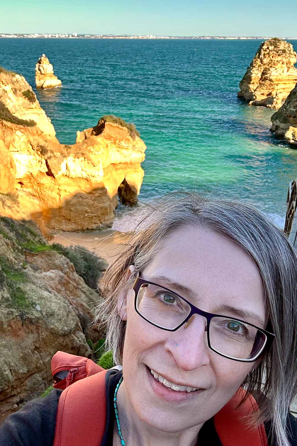 Selfie of smiling woman wearing glasses with turquoise sea and rock formations in background.