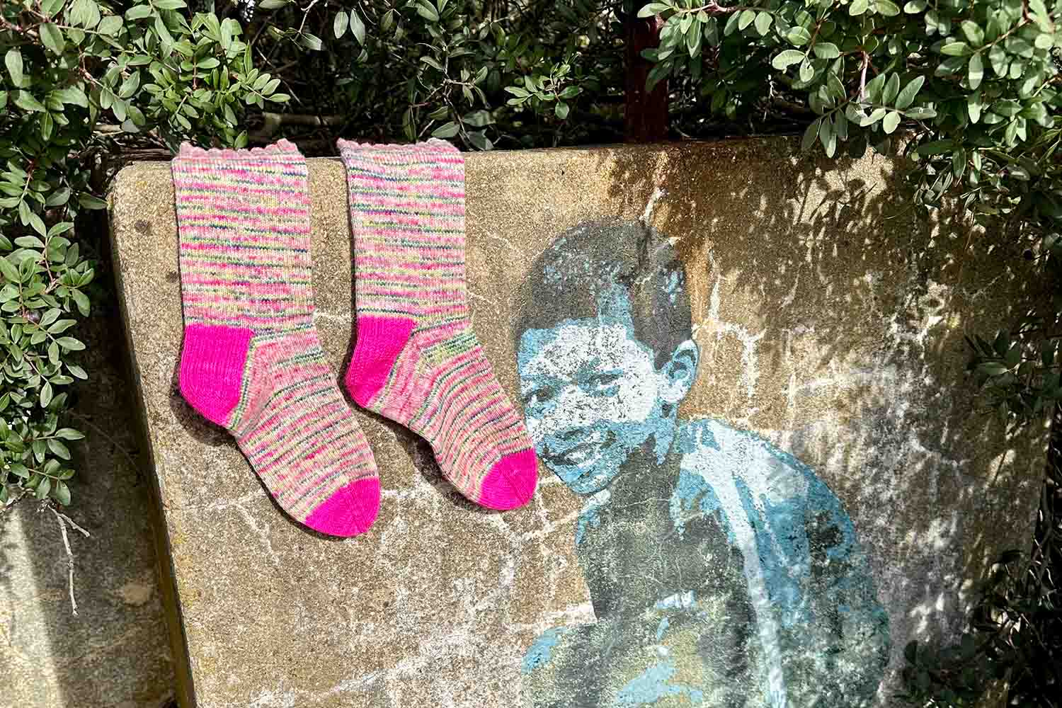 A pair of handknit socks draped over short concrete wall with graffiti art of crouching boy.