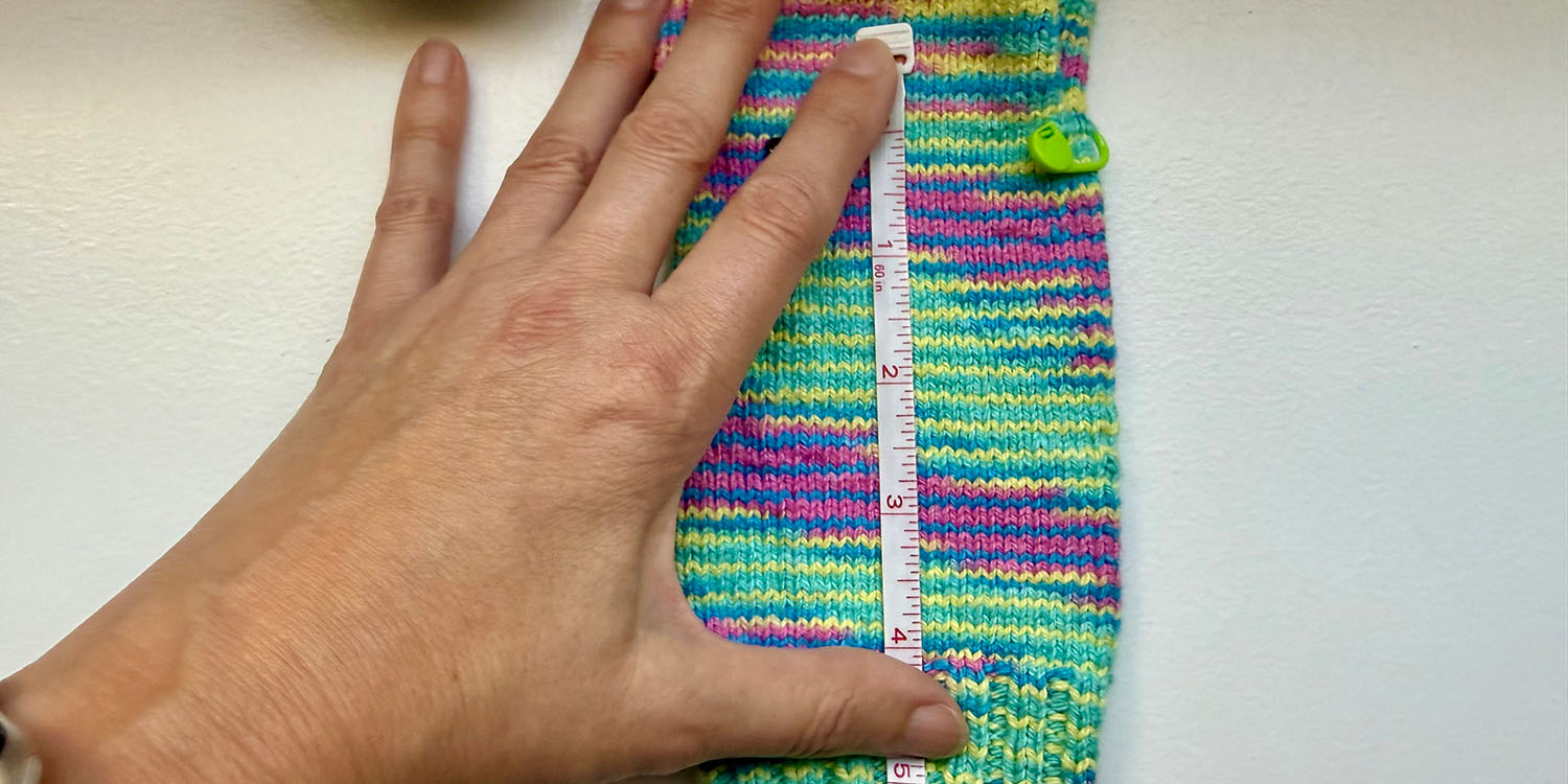Hand holding measuring tape over knitted sock measuring row gauge.